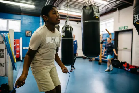 Empire Fighting Chance A male child is captured jumping ropes, he wears a Puma shirt in beige colour and similar colour shorts. In the background we can see another child, also in shorts and a t-shirt but they are blue. They are in a gym and between the two children there are two ceiling mounted punching bags.