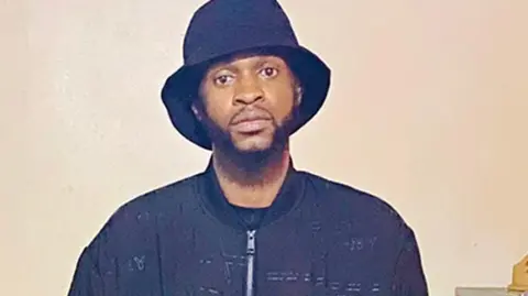 Mvuezolo family Fergany Mvuezolo in a snapshot, wearing a navy zip-up top and a navy hat. He stands in front of a cream-coloured wall.