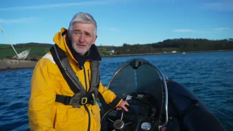 A man standing the wheel of a boat, he has his body facing away from the steering wheel with one hand on it, he is wearing a yellow jacket with grey life vest, he has white hair and beard.
