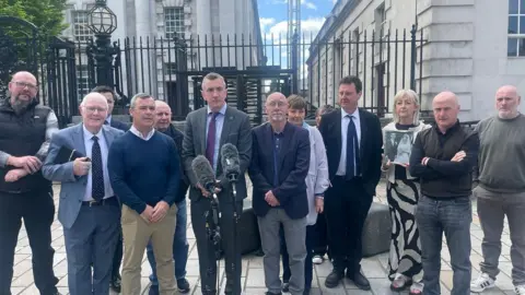 The photo shows nine men and three women standing beside a solicitor dressed in a dark suit outside a court in Belfast. Microphones are in front of the solicitor