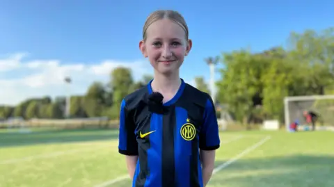 Lara is wearing a Inter Milan shirt. Her hands are behind her back and behind a football pitch.