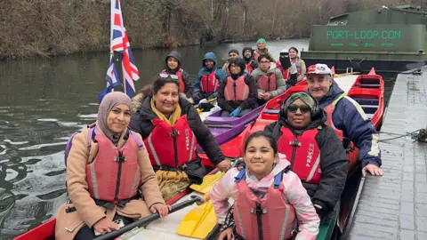 BBC Fourteen people on two boats on a canal in Birmingham. There are 10 women and four men from different ethnicities wearing red life jackets. The boats are parked up by the canal footpath - one at the front is red and the other is purple at the back. There is also a Union Jack flag at the front of the purple boat.