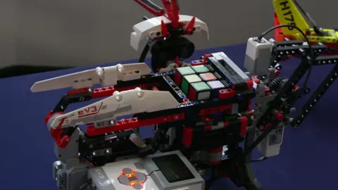 A white and red robot holding a rubik's cube. It is stood on a blue table. 