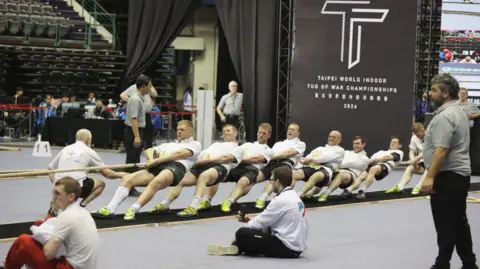 Christine Yang / Chinese Taipei Tug of War Association A tug of war team, all in white shirts and black shorts, strain as they pull on a rope. A scattering of people, seemingly mainly officials are in the background