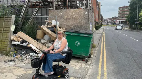 Tony Fisher/BBC Lynda Bell sat on a mobility scooter in front of the scaffolding. She is right next to a road with a car travelling on it. She has blonde hair, glasses and is wearing a white top with blue jeans.