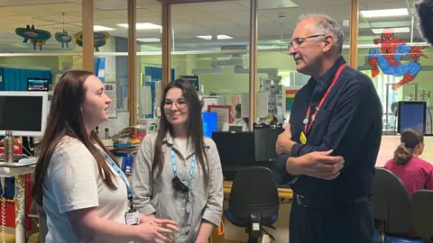 Dr Viktoriya Betsal, Dr Anastasiia Sabadosh and Martin Gargan inside Bristol Royal Hospital for Children. Dr Betsal has long dark red hair and is looking directly at Mr Gargan, having a conversation with him. Dr Sabadosh has long dark brown hair and is looking at Dr Betsal. Mr Gargan is standing with his arms crossed.