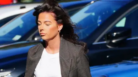 PA Media Ruth walking to court with three cars behind her. She is not looking at the camera. Her hair is blowing in the wind. She is wearing a white t shirt and a blzaer.