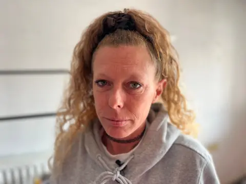 A woman stood in front of a white wall and looking directly at the camera. She is wearing a grey hooded jumper. 