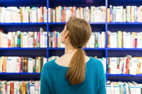 "Rear view of a person with long brown hair in a ponytail, looking at bookshelves filled with books on vibrant blue shelves."