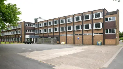 The brick buidling has three floors and two blocks interlinked. There are many uniformed white windows along each floor. The block on the right is boarded up on the first floor. A concrete car park sits in front of the building.