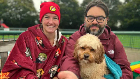 Emma Baugh/BBC Laura Roe, smiling and wearing a red beanie hat, with a maroon-coloured towel around her, sitting next to a man holding a beige dog on his lap