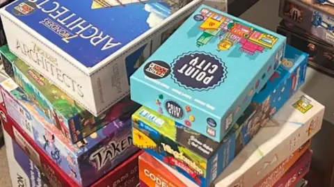 A stack of board games in different shaped and colour boxes. On top is a copy of Point City next to a copy of 6 Wonders Architects