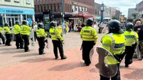 BBC Police officers in a line, wearing riot gear and holding shields, stand near the Blue Clock in Hanley 