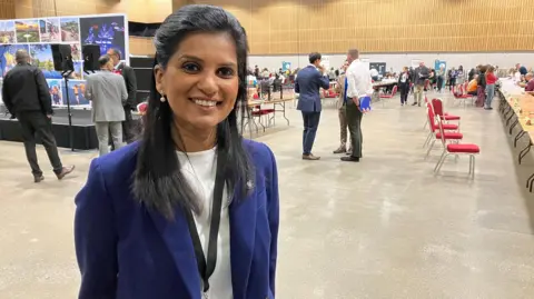 Amy Holmes/BBC Shazna Muzammil smiles at the camera, standing in the election count hall, which has long rows of tables and chairs and a few people milling around.