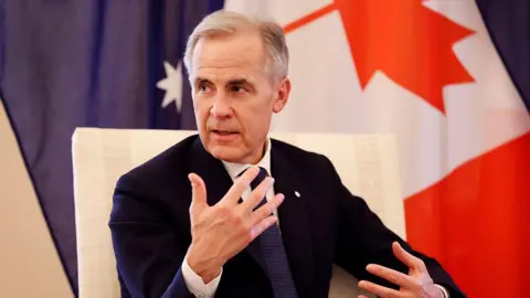 Mark Carney, seated and in a suit and tie, gestures as he speaks