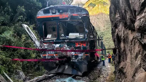 The front of a heavily damaged train carriage stands still on the railway with cordons tied around the area and people in high visibility standing nearby.