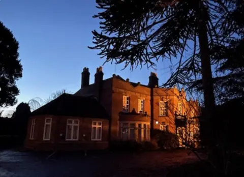 East Staffordshire Borough Council A two-storey building, pictured with light shining on part of the building that is protruding from the rest of the building on that side. A tree, which is dark on the image, is prominent in the foreground on the right.