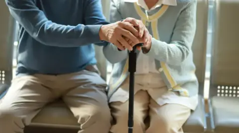 A mid shot of two elderly people. They are both sitting on chairs and holding hands and a walking stick. The person on the left is wearing a blue jumper and light trousers. The person on the right is wearing a light blue cardigan and light trousers.