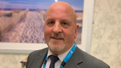 A close-up photo of a man in a grey suit, pink and white striped shirt, and dark blue tie. He is bald with a grey goatee. 