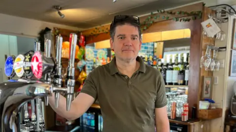 Sara Smith / BBC A man holding a beer tap in a pub behind the bar.
