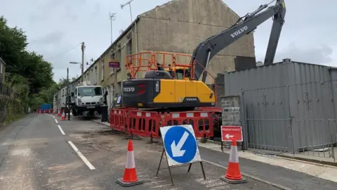 Demolition work on homes in Cyfyng Road, with a digger close to houses and cones and an arrow sign in the street