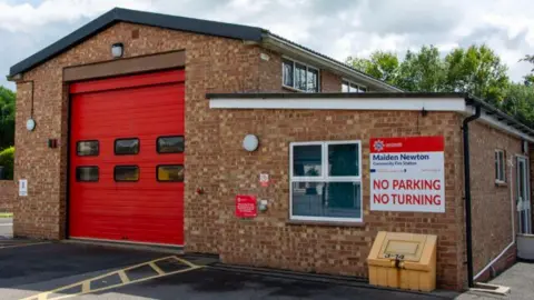 A typical fire station building with a large red garage-style door for vehicles. A sign to the right of the building reads "Maiden Newton Community Fire Station". Underneath that the sign reads "NO PARKING" and "NO TURNING". 