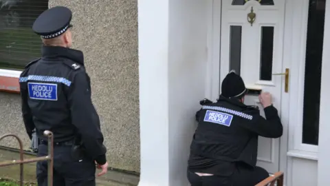 Two police officers standing close to the white front door of a pebbledashed house in Bangor, both wearing black uniforms.  One in a flat hat looks on as his colleague wearing a traditional helmet opens the letterbox to speak to people inside and ask them to open the door