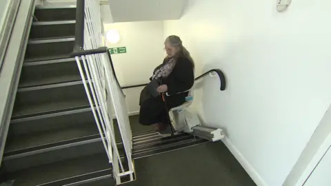 The image shows the stairwell of some flats, with a stairlift. Lynda Wilkins is sitting in the stairlift holding a walking stick.