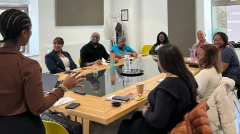 Our Version Media Eight people sitting around a large wooden table during a course. Course instructor and Our Version Media founder Veronica Gordon is standing at the front, teaching. The participants face Veronica. There are glasses of water on the table.