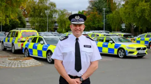 Cambridgeshire Police Simon wearing white police shirt and black trousers and a black tie and Black police hat- standing in front of police cars.