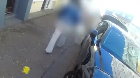 The view from a civil enforcement officer's body-worn camera which shows a parking ticket lying on the pavement as a woman walks away from it past a parked car. The image of the woman is blurred.