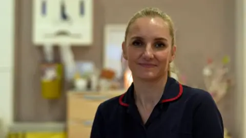 Tabitha Stuthridge is smiling at the camera, in a dark blue uniform, and has blonde hair tied back. She is sitting in a hospital setting.