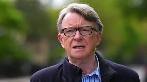 Lord Mandelson in glasses and a blue striped shirt 
