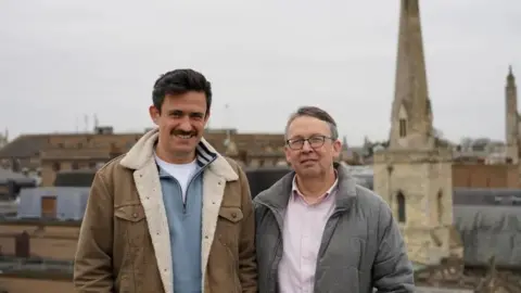 Peter Richer (left) with short dark hair and moustache, wearing a brown coat, grey sweater and white T-shirt, and Mark Richer with short dark hair and glasses, wearing a grey coat and light-coloured shirt. There is a church spire behind them in light stone and other buildings are visible.