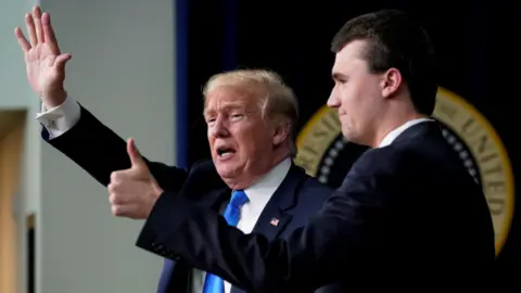 Reuters Charlie Kirk gives a thumbs-up as he stands in profile next to Donald Trump who is waving. Both are wearing suits.