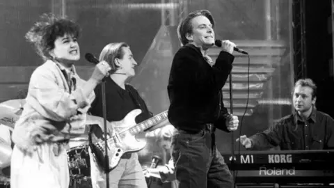 A black and white photo of a Deacon Blue performance. Lorraine McIntosh and Ricky Ross are singing into a mics  and there is also a guitarist and keyboard player in the picture.