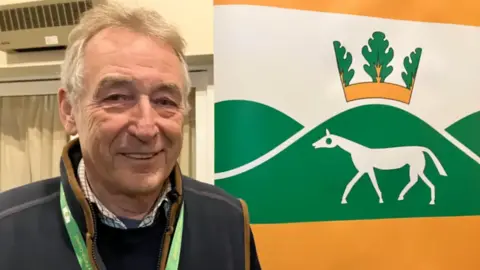 A man is standing with a sign behind him which has a green hill with a chalk horse on it. The man is smiling at the camera and is wearing a jacket with checked shirt. He is also wearing a lanyard. There is a patio door behind him.