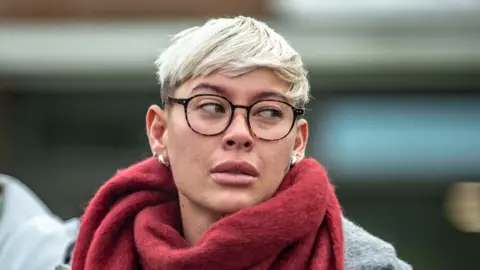 Andrew Price A woman with short blonde hair and glasses, wearing a grey court and red scarf