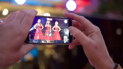 A man holding up a phone to record three singers in red dresses
