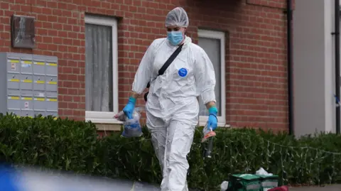 PA Media A forensic officer walks on a pavement carrying evidence bags behind a police cordon. A block of flats is behind them with two green first aid bags also sat on the pavement outside the block.