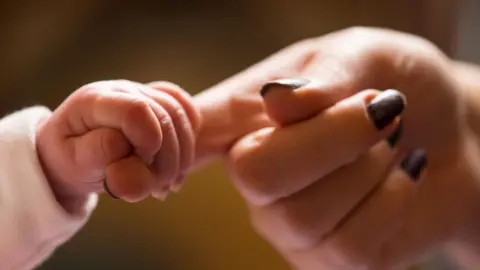PA Media A baby's hand is holding a woman's index finger.