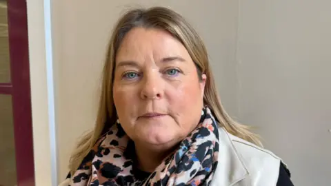 Shows a woman with fair hair and blue eyes looking at the camera. She is wearing a colourful scarf and light grey jacket with black sleeves. She is standing in front of a wall.