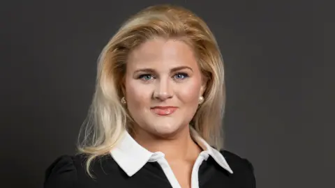 Alex Davies-Jones is smiling in the head and shoulders picture. She has-shoulder length blonde hair which falls on a black top with a large white collar. She is wearing large pearl earrings.