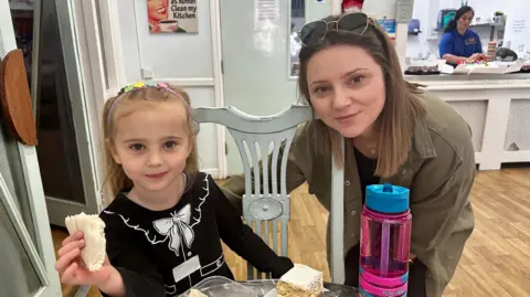 A woman and her four-year-old daughter are looking staright to the camera. The little girl is smiling and has a sandwich in her hand.