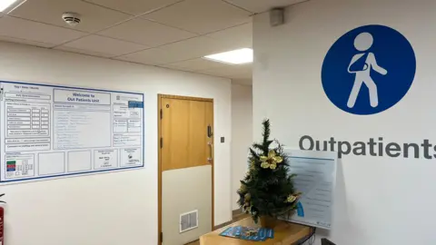 The Outpatient unit at the Princess Elizabeth Hospital. There is a blue symbol of a person with a sling on their arm on the white wall. A small Christmas tree is placed on the reception desk, with gold flowers on it. There are a few leaflets next to the tree. A welcome board is on the wall next to a brown cupboard door. 