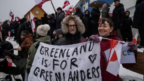 Reuters People attend a protest against U.S. President Donald Trump's demand that the Arctic island be ceded to the U.S., calling for it to be allowed to determine its own future, in front of the U.S. consulate in Nuuk, Greenland, January 17, 2026. They're holding a banner reading, Greenland is for Greenlanders
