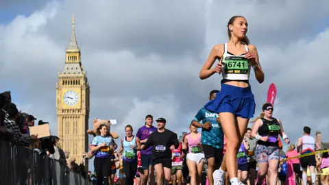 A large group of people participate in the London Landmarks Half Marathon, running on a city street with the Elizabeth Tower visible in the background under a cloudy sky.