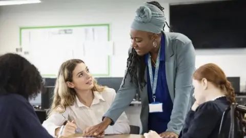 A teacher stands beside a classroom desk, helping pupils