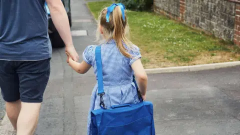 A little girl with a blue school dress and with a large blue bag, walks down the pavement hand-in-hand with an adult.