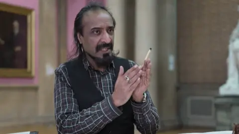 A smartly dressed man talking while holding a stylus. He appears to be sat in a large hall in a gallery or museum. 